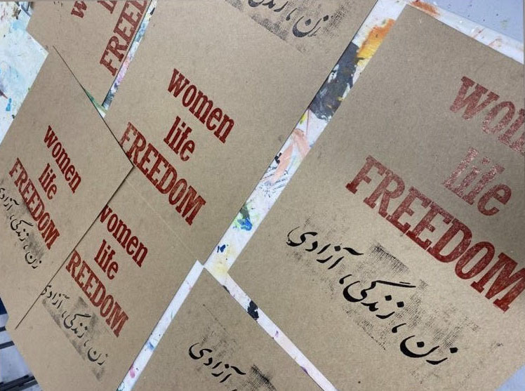 Multiple letterpress printed posters laid on a table printed on brown cardstock with slogan "Women, Life Freedom" printed in red ink in English and black ink in Farsi.