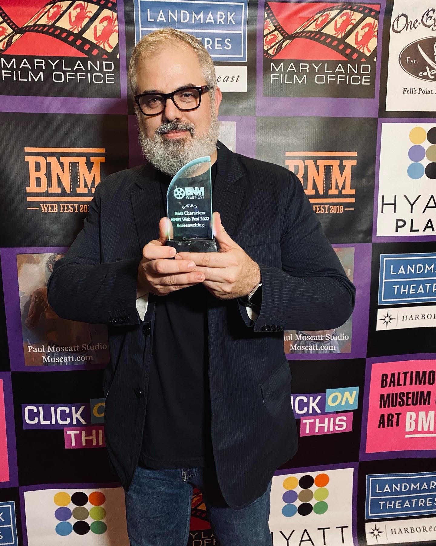 A photo of Joseph Ross Angelella holding an award standing in front of a step and repeat backdrop