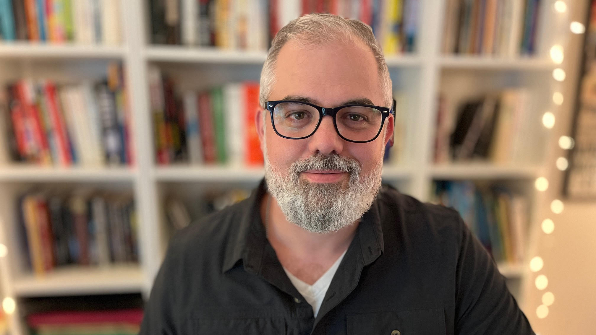 Photo of bearded man wearing glasses in front of bookshelf