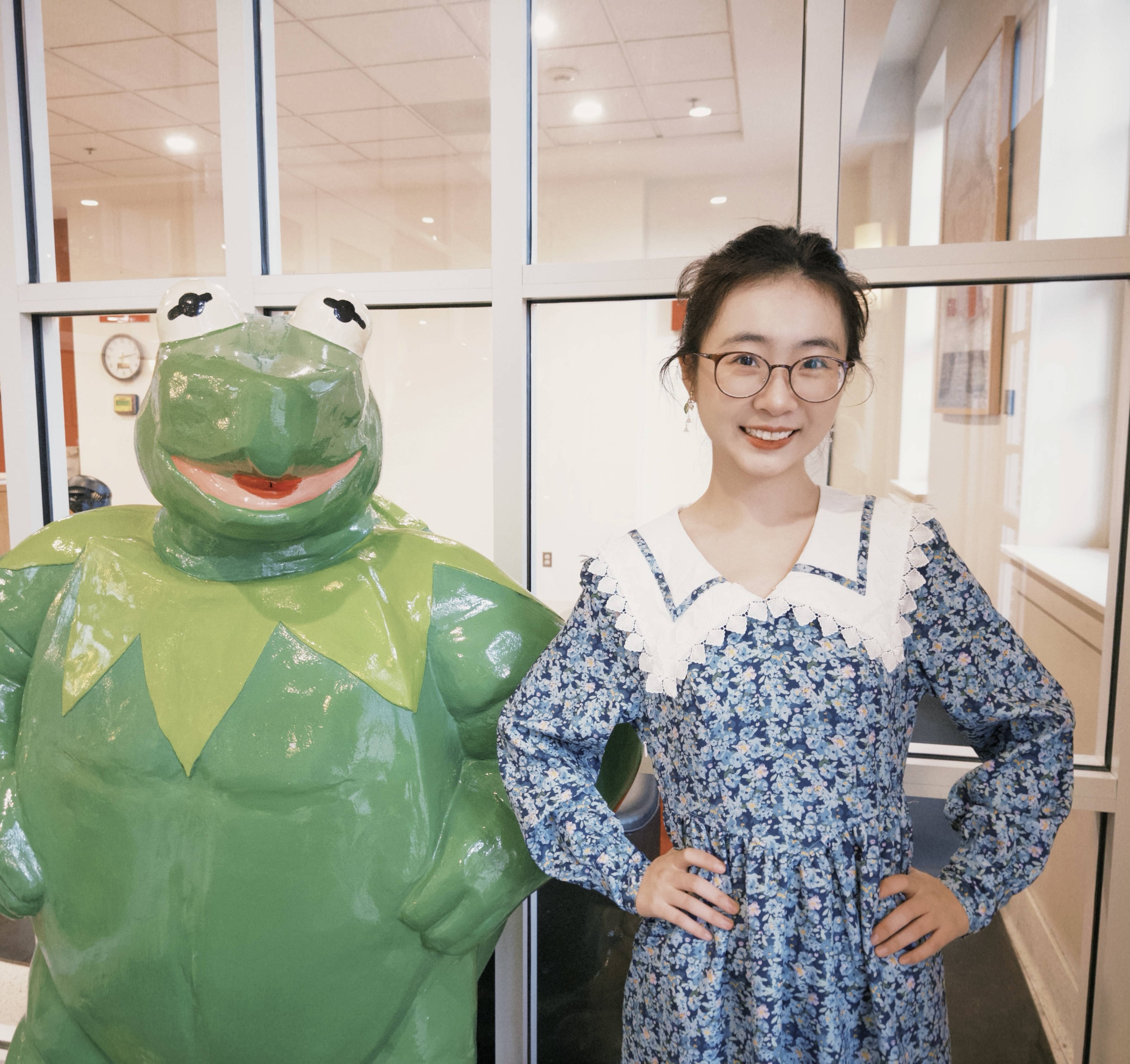 Smiling woman standing, hands on hips, next to Kermit the Frog statue in same pose