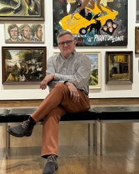 A man sits on a bench and looks at the camera. He is surrounded by colorful pieces of art.