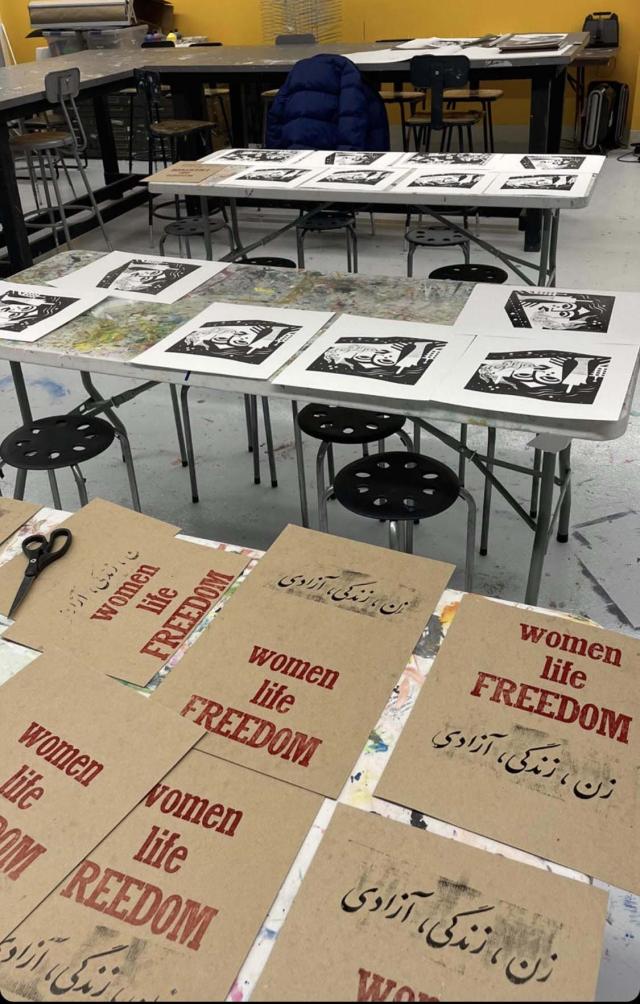 Multiple letterpress posters printed on brown cardstock with slogan "Women, Life Freedom" printed in red ink in English and black ink in Farsi, laid on a table. On another table lay many screen prints in black ink with an illustration of an Iranian woman protesting.